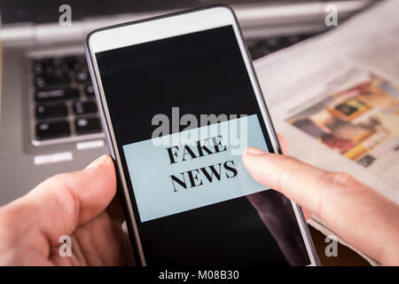 La main de l'homme tenant un smartphone avec Fake News mots sur l'écran sur un journal et un ordinateur portable. Canular et Fake news concept. Banque D'Images