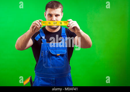 Un jeune homme builder worker wearing un t-shirt noir et bleu dans l'ensemble de la construction utilise de niveau magnétique rare sur un fond vert isolés Banque D'Images