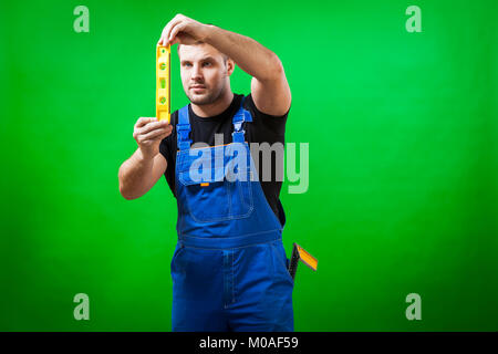 Un homme aux cheveux noirs construction worker wearing un T-shirt noir et bleu dans l'ensemble de la construction utilise de niveau magnétique règle sur un vert isolés backgroun Banque D'Images