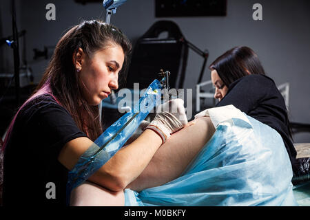 Jeune femme maître de tatouage tatouage tatouage fait avec machine à sous l'ampoule lumineuse sur la hanche de belle femme dans l'atelier sombre, vue de l'arrière Banque D'Images