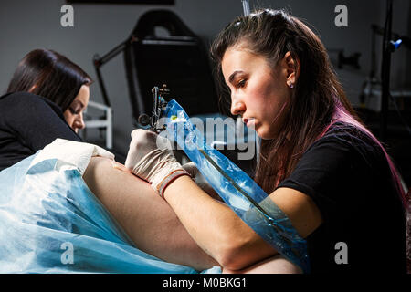 Jeune femme maître de tatouage tatouage tatouage fait avec machine à sous l'ampoule lumineuse sur la hanche de belle femme dans l'atelier sombre, vue de l'arrière Banque D'Images