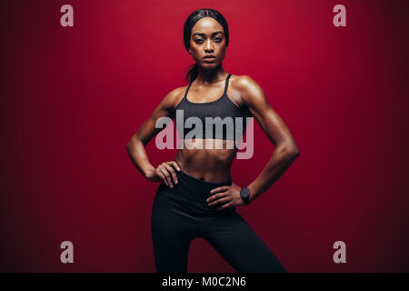 Young woman wearing sportswear noir debout contre l'arrière-plan rouge. Femme africaine en bonne santé avec un corps musclé standing in studio. Banque D'Images