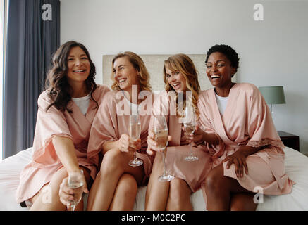 Belle et heureuse mariée demoiselles célébrant hen party avec champagne dans la chambre. Les femmes en robe à dormir assis sur le lit et avoir du champagne. Banque D'Images