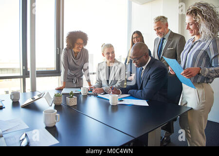 Groupe diversifié de businesspeople standing autour de leur directeur assis à une table de réunion du bureau de l'examen des graphiques et de la paperasserie Banque D'Images