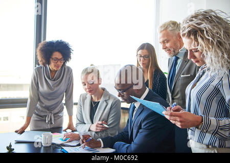 Groupe diversifié de l'accent businesspeople standing autour de leur directeur assis à une table de réunion du bureau de l'examen des graphiques et de la paperasserie Banque D'Images