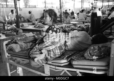 Une femme berceaux son bébé assis sur le lit de son mari gravement malade dans l'hospitalisation à l'unité clinique Mae Tao sur la frontière thaïlando-birmane. Environ 300 personnes par jour reçoivent des soins gratuits à la clinique, la plupart étant des travailleurs migrants et réfugiés qui ont franchi la frontière de la Birmanie (Myanmar) pour échapper à la pauvreté désespérée et la persécution du régime militaire. Les soins de santé sont tous mais inexistante en Birmanie, où moins de 2  % du PIB est consacré à la santé et l'éducation combinées. Banque D'Images