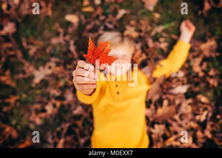 Garçon couché dans les feuilles d'automne holding up a leaf Banque D'Images