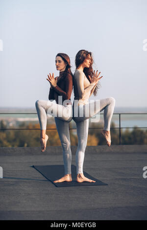 In l'article couple yoga pose dos à dos sur le toit à l'extérieur. Deux belles femmes se concentrent sur faire de l'exercice tout en se tenant par l'augmenter Banque D'Images