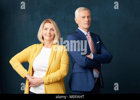 Studio shot of senior couple debout à fond sombre. Smiling mature woman with hand sur sa hanche à la caméra et à un homme âgé portant fonction whi Banque D'Images