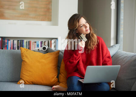 Une femme assise sur un canapé et réfléchis à la pendant leurs achats en ligne avec sa carte credeit. Banque D'Images