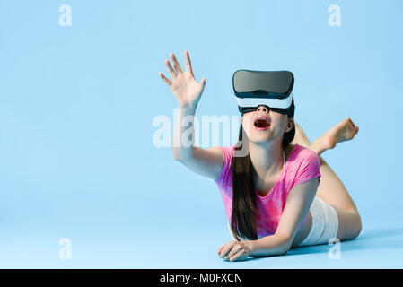 Belle jeune fille couchée sur fond bleu marbre en situation périphérique et à l'aide de la technologie VR de toucher la main de l'écran de simulation sentiment excité. Banque D'Images