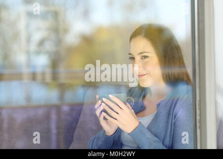 Femme sérieuse à la recherche de l'avant par une fenêtre à la maison avec des réflexions sur la vitre Banque D'Images