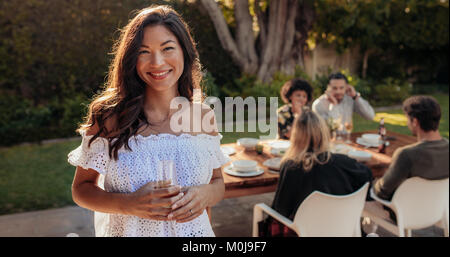 Belle asiatique femme debout en plein air avec des amis assis en arrière-plan ayant la nourriture. Femme avec un verre au partie en plein air. Banque D'Images