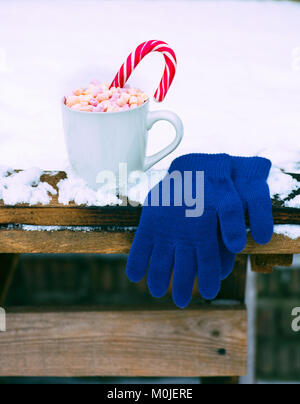 Une coupe grey avec du chocolat chaud et de la guimauve sur une table couverte de neige dans la rue, à côté d'une paire de mitaines en tricot bleu, une journée d'hiver Banque D'Images