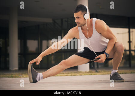 Young sportsman doing stretching exercice, la préparation de la formation dans le parc. Tout en s'exerçant, il écoute de la musique via heasphones. Fitness, le sport, les natures mortes Banque D'Images