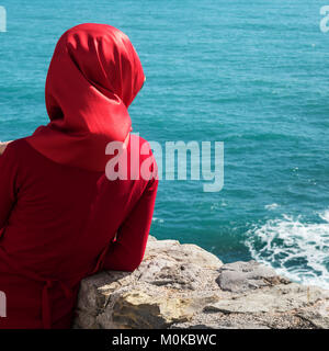 Une femme portant un foulard rouge et robe rouge se tient à un mur de pierre en voyant l'eau bleue, Budva, Budva, Monténégro Municipalité Banque D'Images