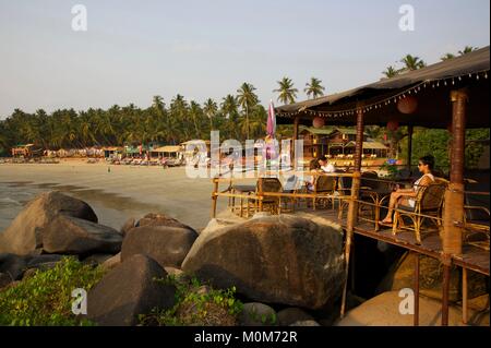 L'Inde, Goa, Palolem, les touristes d'une terrasse d'un bar donnant sur une plage de sable doré bordée de restaurants et boutiques, entre les rochers et arbres cocnut Banque D'Images