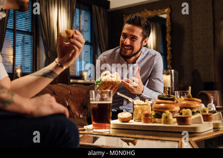 L'homme est de rire et de parler à son ami autour d'un repas dans un bar/restaurant. Ils mangent des hamburgers et boire de la bière. Banque D'Images