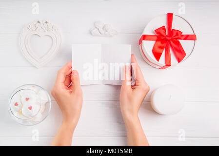 Women's Hands holding empty carte de souhaits sur fond blanc avec une décoration romantique comme cadre, coffret en forme de coeur, anges, statuette, des bonbons . Le jour de la Saint-Valentin. Banque D'Images