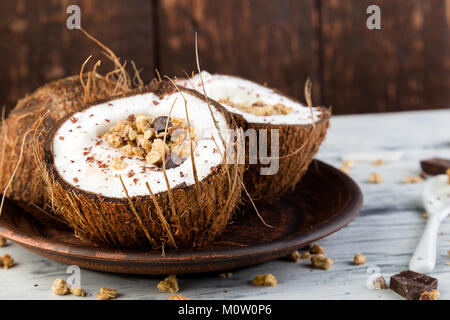 Petit-déjeuner sain dans la noix de coco noeud sur fond blanc. Le yogourt dans un bol de noix de coco avec des flocons de noix de coco, chocolat et muesli. Vue de dessus, des laïcs, des frais généraux Banque D'Images