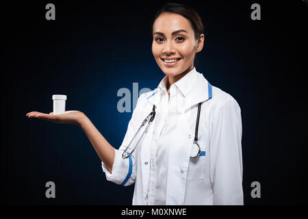 Jeune brunette infirmière en blouse médicale montrant une bouteille de pilules et à la plus isolée de l'appareil photo noir Banque D'Images