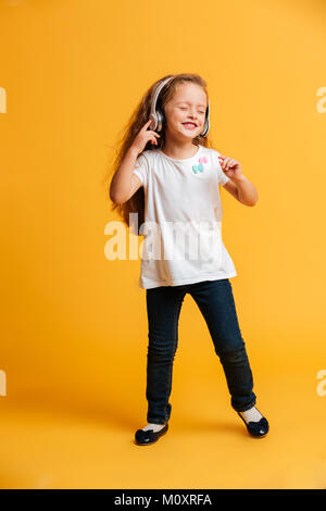 Photo de petite fille isolée sur fond jaune à danser à l'écoute de la musique avec des écouteurs. Les yeux fermés. Banque D'Images