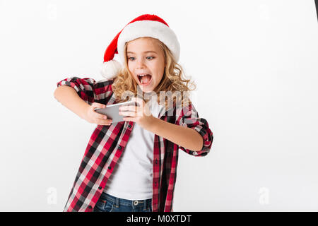 Portrait d'une petite fille excitée vêtue de christmas hat jouer aux jeux sur téléphone mobile isolé sur fond blanc Banque D'Images