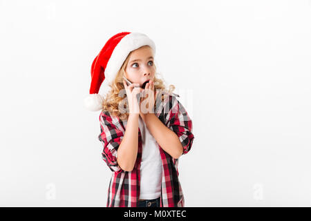 Portrait d'une petite fille surprise habillé de christmas hat talking on mobile phone while looking at copie espace isolé sur fond blanc Banque D'Images