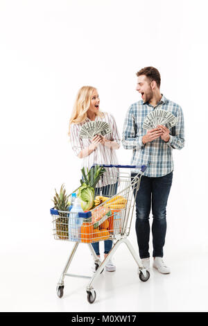Portrait d'un couple heureux holding tas de billets et de l'argent à l'un l'autre en se tenant debout avec un chariot de supermarché o isolés Banque D'Images