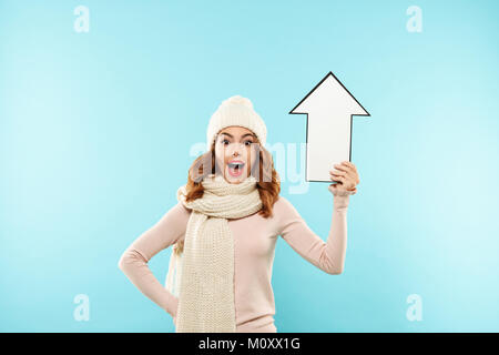 Portrait of a happy smiling girl in winter clothes holding paper-flèche haut isolé sur fond bleu Banque D'Images