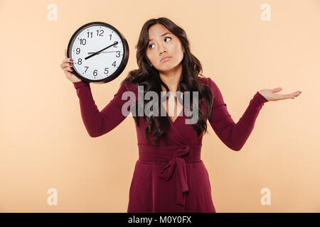 Femme brunette perplexe avec de longs cheveux bouclés holding horloge indiquant l'heure après 8, faisant des gestes comme si elle est en retard ou ne se soucient pas sur fond de pêche Banque D'Images