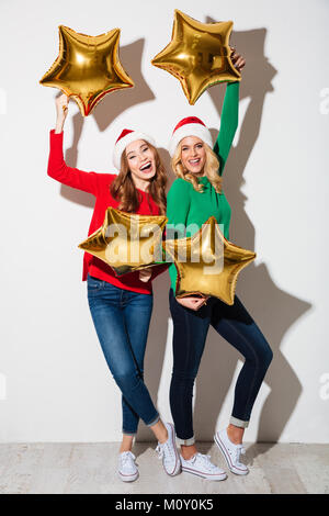 Portrait de deux jeunes filles gaies vêtus de chandails et casquettes noël posing while holding montgolfières et isolé de ove Banque D'Images