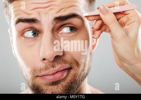 Close up portrait of female part plumer les sourcils de jeunes hommes avec des brucelles isolées sur fond gris Banque D'Images