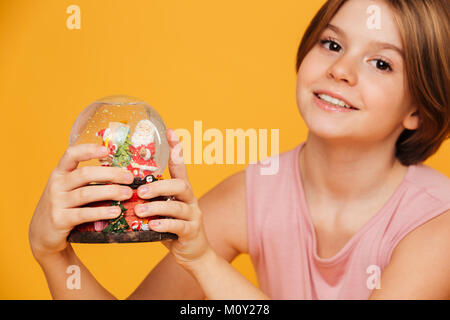 Portrait of beautiful smiling girl en robe rose à la caméra et holding globe de neige dans les mains sur jaune isolé Banque D'Images