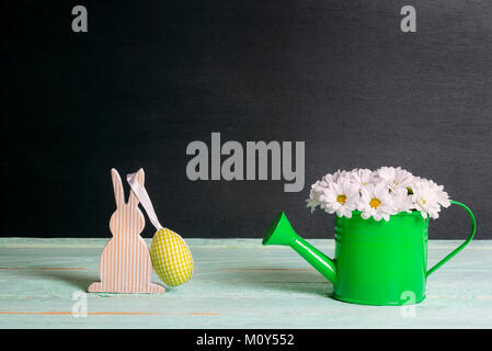 Cute white daisies arrangement dans un arrosoir et un lapin en bois avec un tissu jaune egg hanging, sur un tableau vert et un fond noir. Banque D'Images