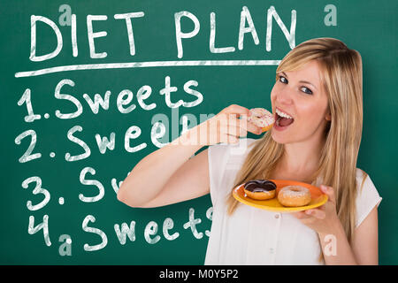 Young Woman Eating Donuts standing in front of Blackboard avec écrit dessus plan de régime Banque D'Images