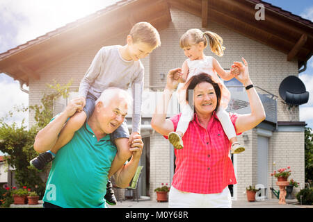 Smiling petits-enfants est assise sur l'épaule de grand-parent bénéficiant à l'extérieur de leur maison Banque D'Images