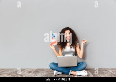 Portrait de femme satisfaite avec beau sourire assis en lotus posent avec l'ordinateur d'argent sur les jambes, le holding american flag dans la main sur mur gris Banque D'Images