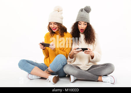 Deux belles filles en pulls et bonnets assis sur le plancher, tandis qu'à l'aide de leurs smartphones sur fond blanc Banque D'Images