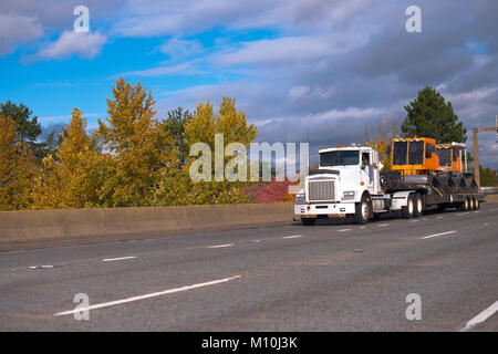 Big Rig semi blanc puissant camion transportant d'orange road de l'équipement de travail véhicules sur l'étape vers le bas semi-remorque déménagement sur une large route d'automne Banque D'Images