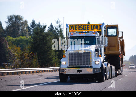 Puissant blanc gros camion semi truck avec charge surdimensionnée signer et descendre effectuer remorque et transportent d'énormes digger surdimensionnée sur autoroute large multiligne Banque D'Images