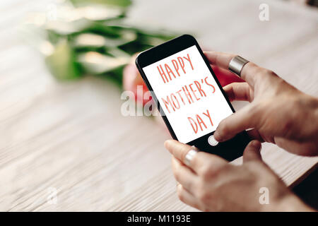 Happy mother's day signe texte à l'écran de téléphone. hand holding gadget avec message ci-dessus des tulipes roses sur fond rustique en bois blanc carte de vœux. Banque D'Images