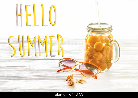 Hello summer texte, vacances. cocktail jaune avec le jus d'abricot, de lunettes et de coquilles en clair sur fond blanc en bois rustique et de voyage. Banque D'Images