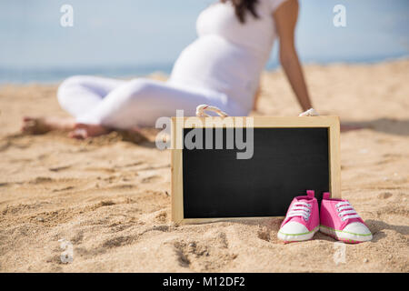 Le portrait d'une paire de chaussures de bébé rose et un mini-tableau sur le sable avec femme enceinte sur l'arrière-plan Banque D'Images