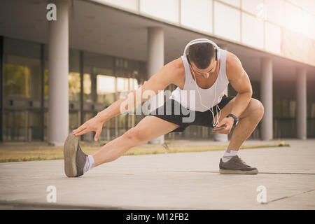 Young sportsman doing stretching exercice, la préparation de la formation dans le parc. Tout en s'exerçant, il écoute de la musique via heasphones. Fitness, le sport, les natures mortes Banque D'Images