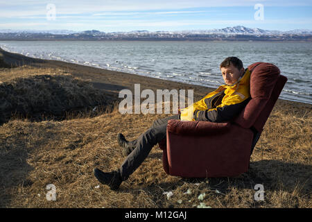 Homme sérieux est assis sur le fauteuil rouge sur la falaise couverte par l'herbe décolorée sur le fond de la mer et montagnes enneigées en Islande. Il regarde dans Banque D'Images