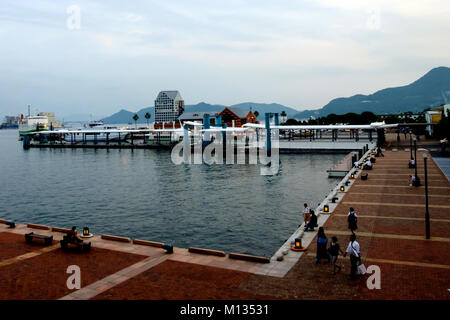 Avis de Sasebo, au Japon par la mer avec des bateaux à quai et les gens se détendre, assis et en marchant, en regardant l'eau. Montagnes en arrière-plan. Banque D'Images