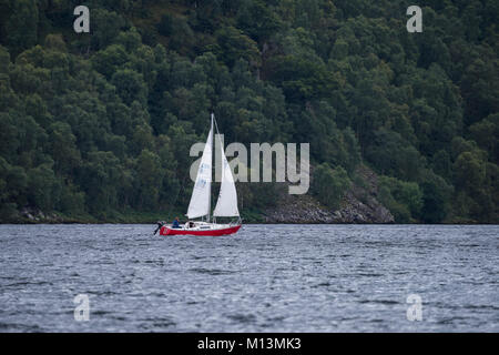 Avec des pentes colline boisée au-delà, un petit bateau rouge avec 2 voiles, navigue sur l'eau du lac Ullswater - Lake District, England, UK. Banque D'Images