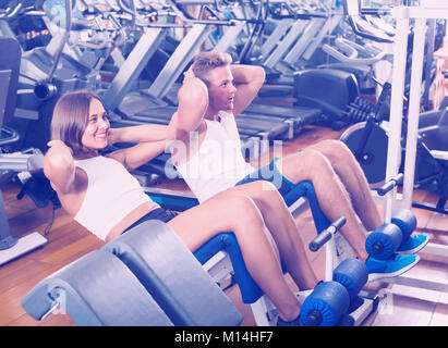 Heureux l'homme et de la femme faisant asseoir ups ensemble à l'aide de la machine en salle de sport Banque D'Images