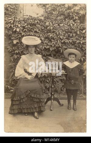 Carte postale Edwardienne originale et charmante de mère et de son petit fils. Ils portent de grands chapeaux à l'extérieur dans le jardin. La femme porte un chemisier de dentelle et un grand chapeau de dentelle. Le jeune garçon porte un costume de marin tendance. Une fougère dans un conteneur se trouve sur la table. Vers. 1903, ROYAUME-UNI Banque D'Images